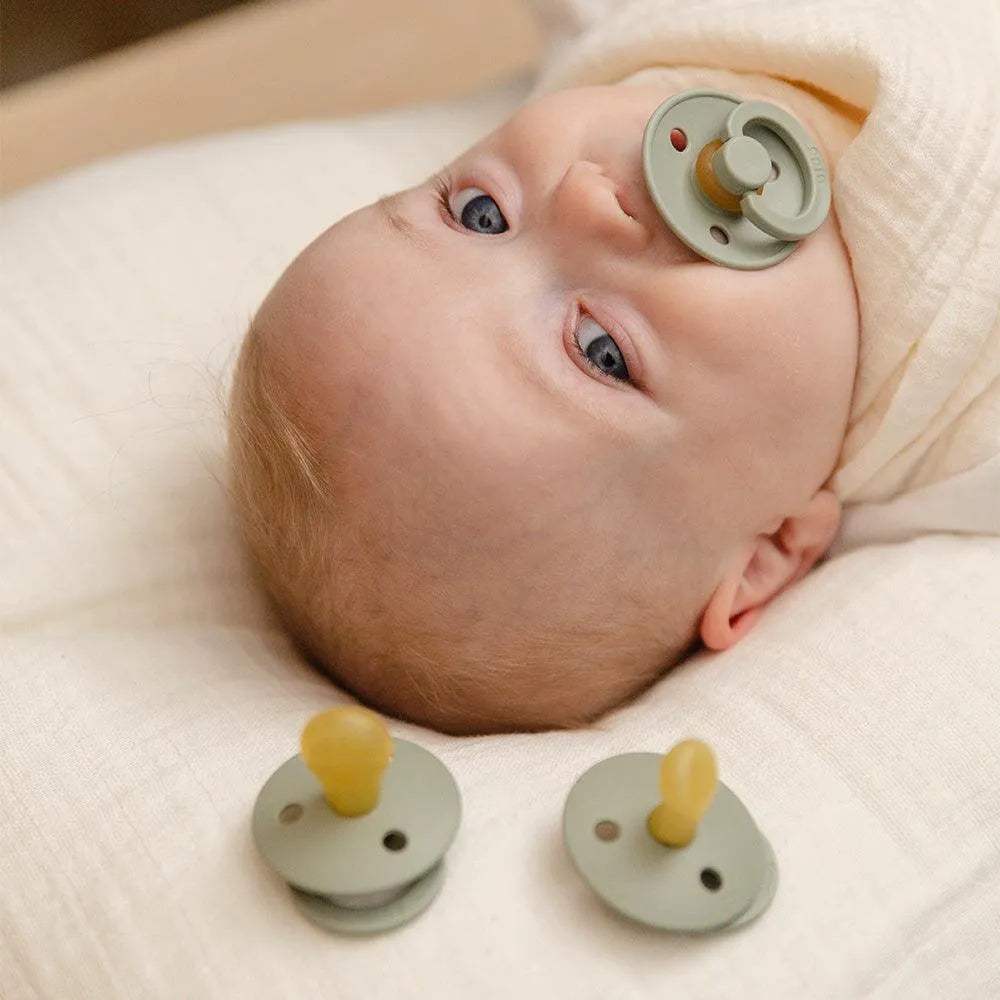Bébé allongé avec une sucette BIBS, deux autres sucettes vert sauge posées devant lui