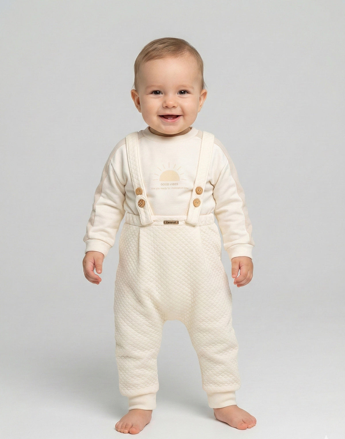 Bébé debout portant l'ensemble Caramell écru, sweat et pantalon matelassé à bretelles